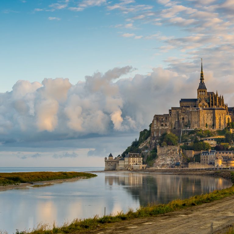 A majestic castle sits atop a rocky island with steep walls, surrounded by calm water. Fluffy clouds and a serene sky create a tranquil, picturesque scene.