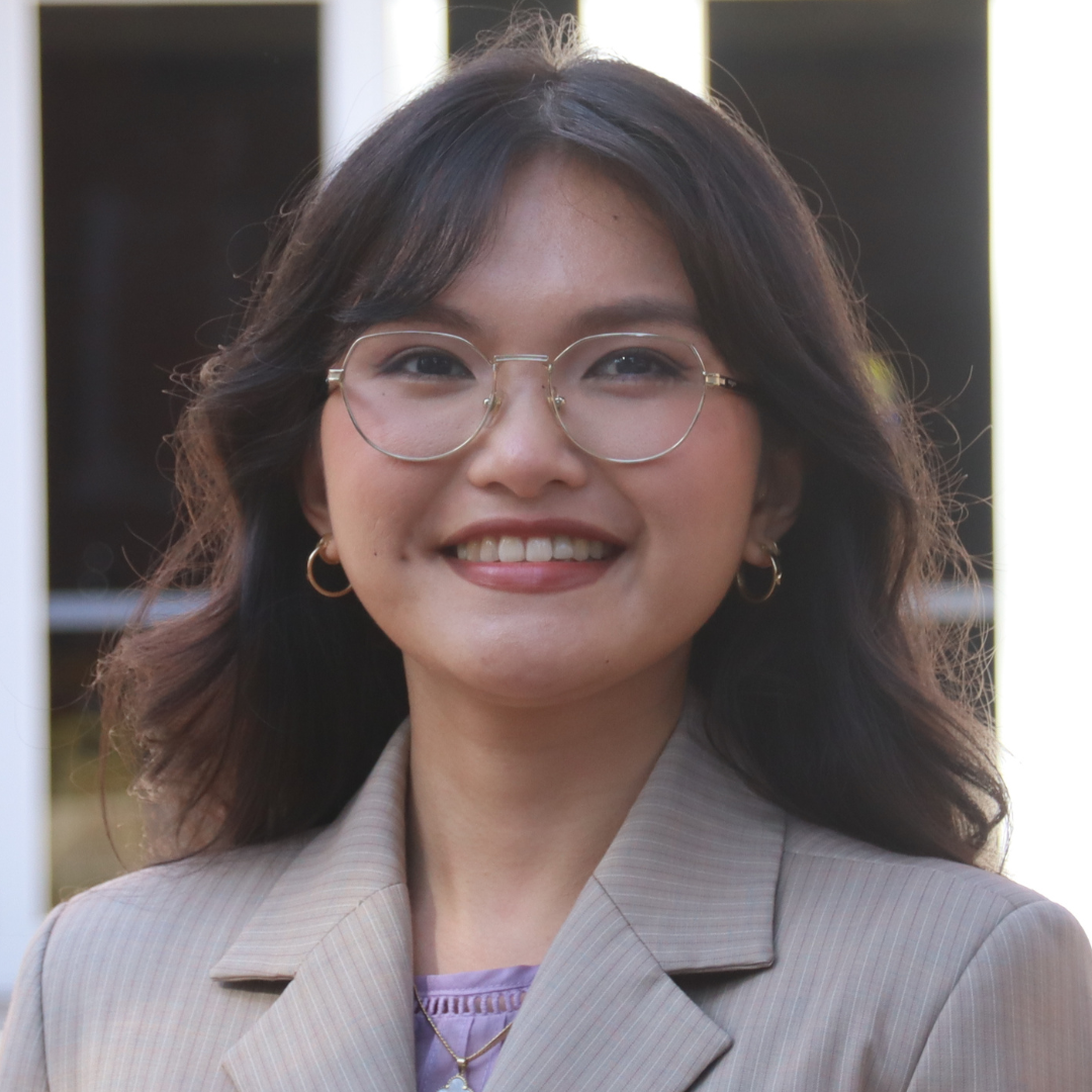 A smiling young woman with dark wavy hair and glasses wears a beige pinstriped blazer over a purple top.