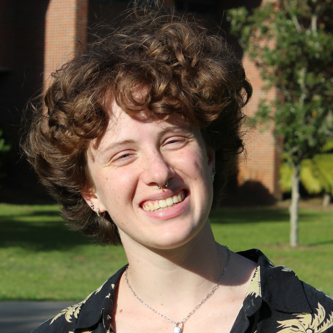 A smiling person with curly brown hair, a septum piercing, and a padlock necklace stands outdoors.