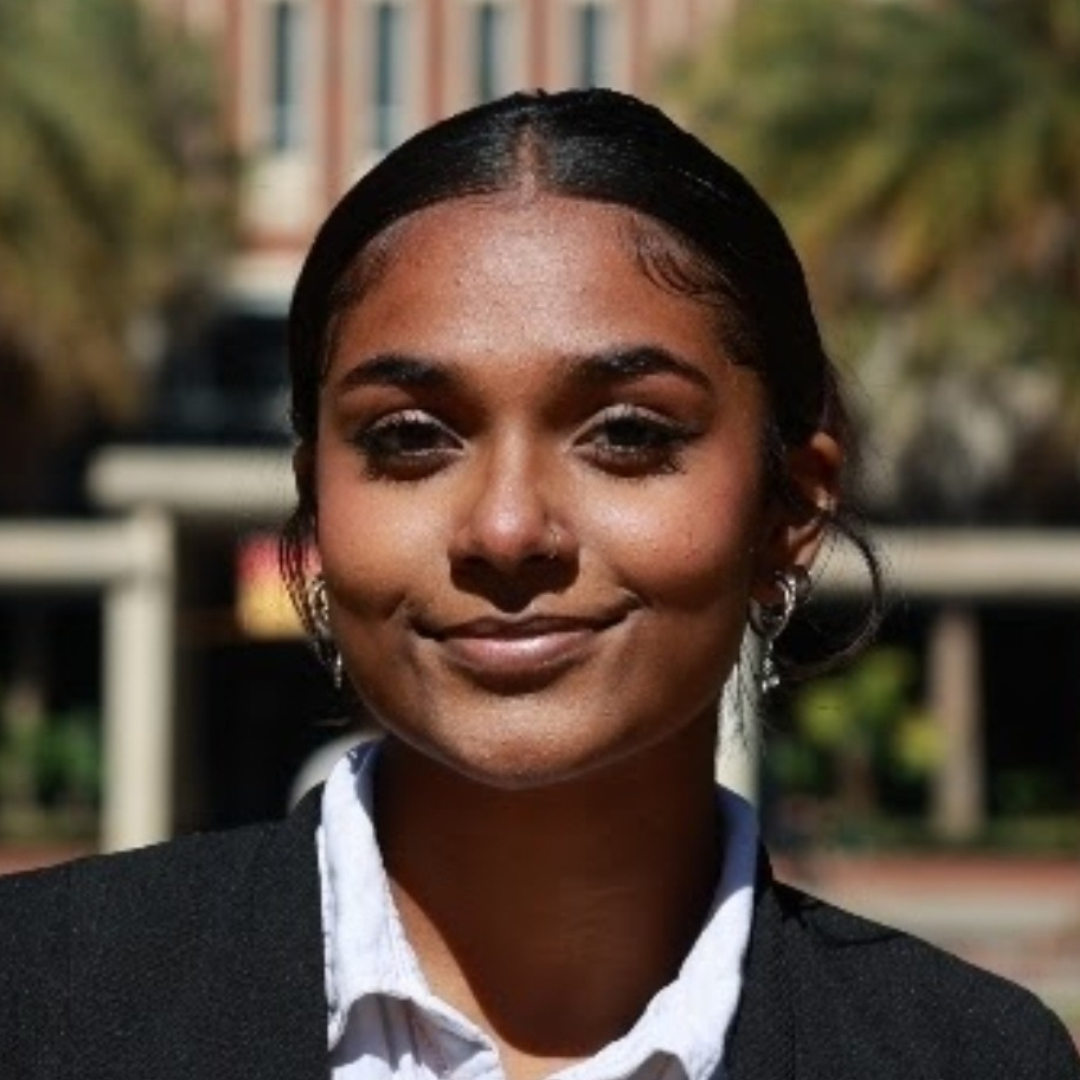 Headshot of a smiling young woman with dark hair and a nose piercing, wearing a blazer over a white shirt, with a blurred outdoor background.