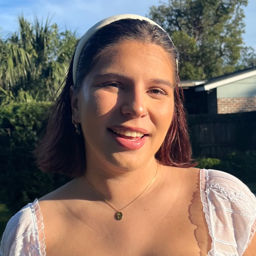 Smiling young woman with a white headband, nose ring, and a gold pendant necklace, illuminated by sunlight outdoors with a blurred background of trees and a house.
