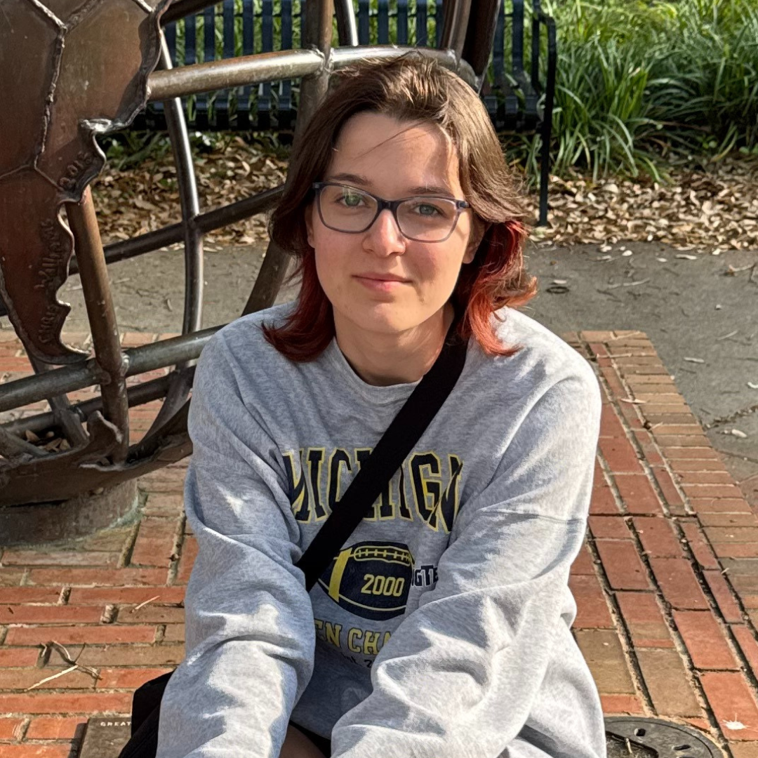 A young person with glasses and brown hair with red streaks smiles, wearing a grey sweatshirt displaying "MICHIGAN", "2000 GT", and "EN CH", sitting outdoors on a brick surface.