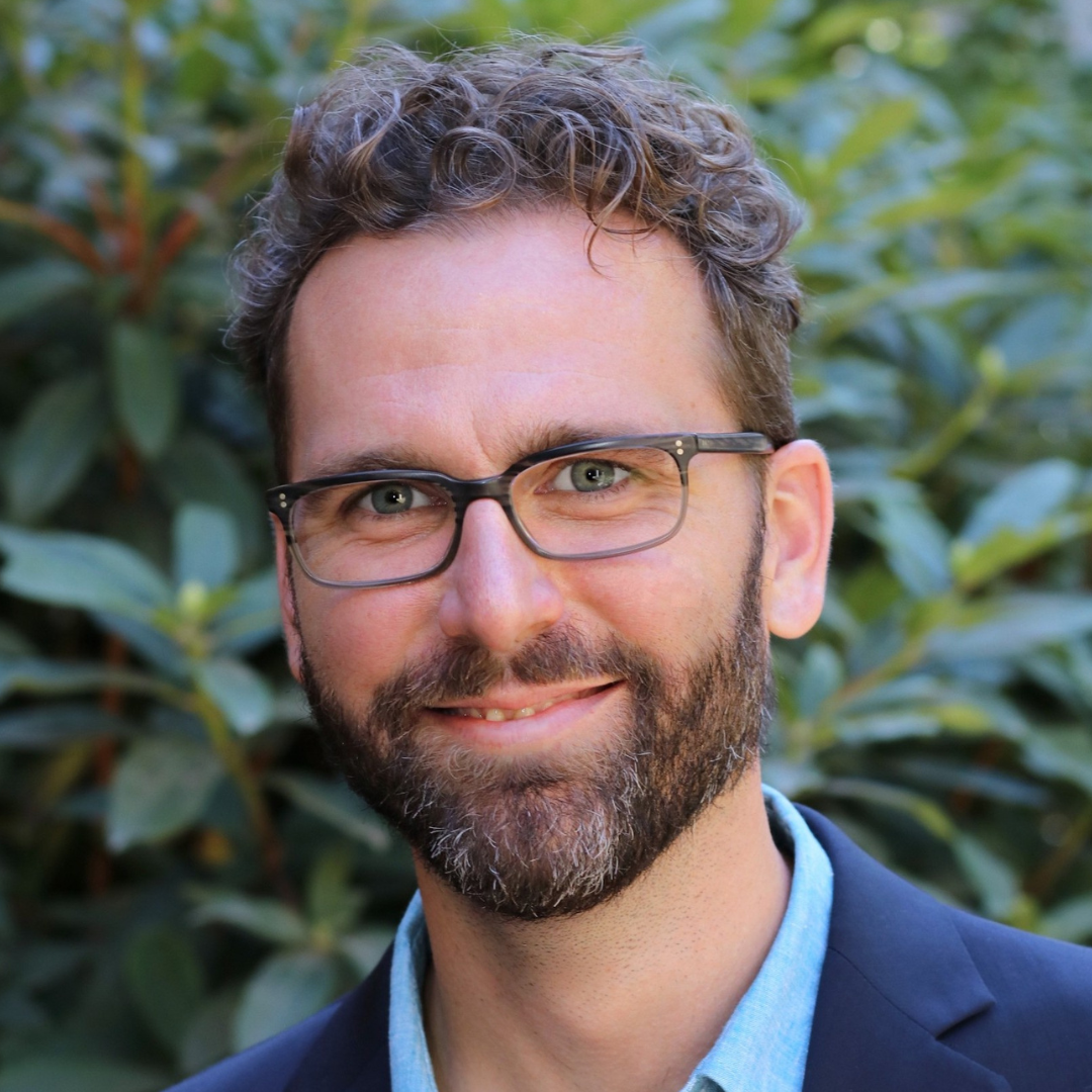 Derek Burdette: A smiling man with curly hair and glasses, wearing a suit jacket and blue shirt. He stands in front of lush green foliage, exuding a friendly and professional demeanor.