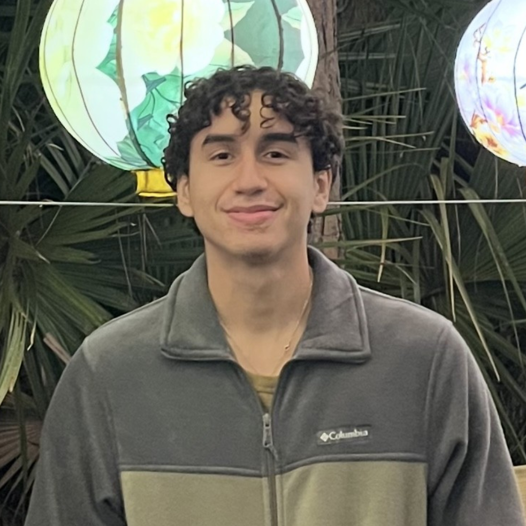 Smiling young man wearing a gray and olive green Columbia fleece jacket and a gold chain, standing outdoors in front of decorative lanterns and palm leaves.