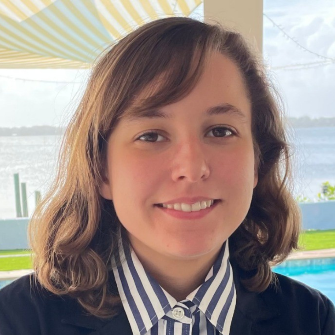 Young woman smiling, wearing a navy blazer and striped shirt, with a body of water and pool in the background.