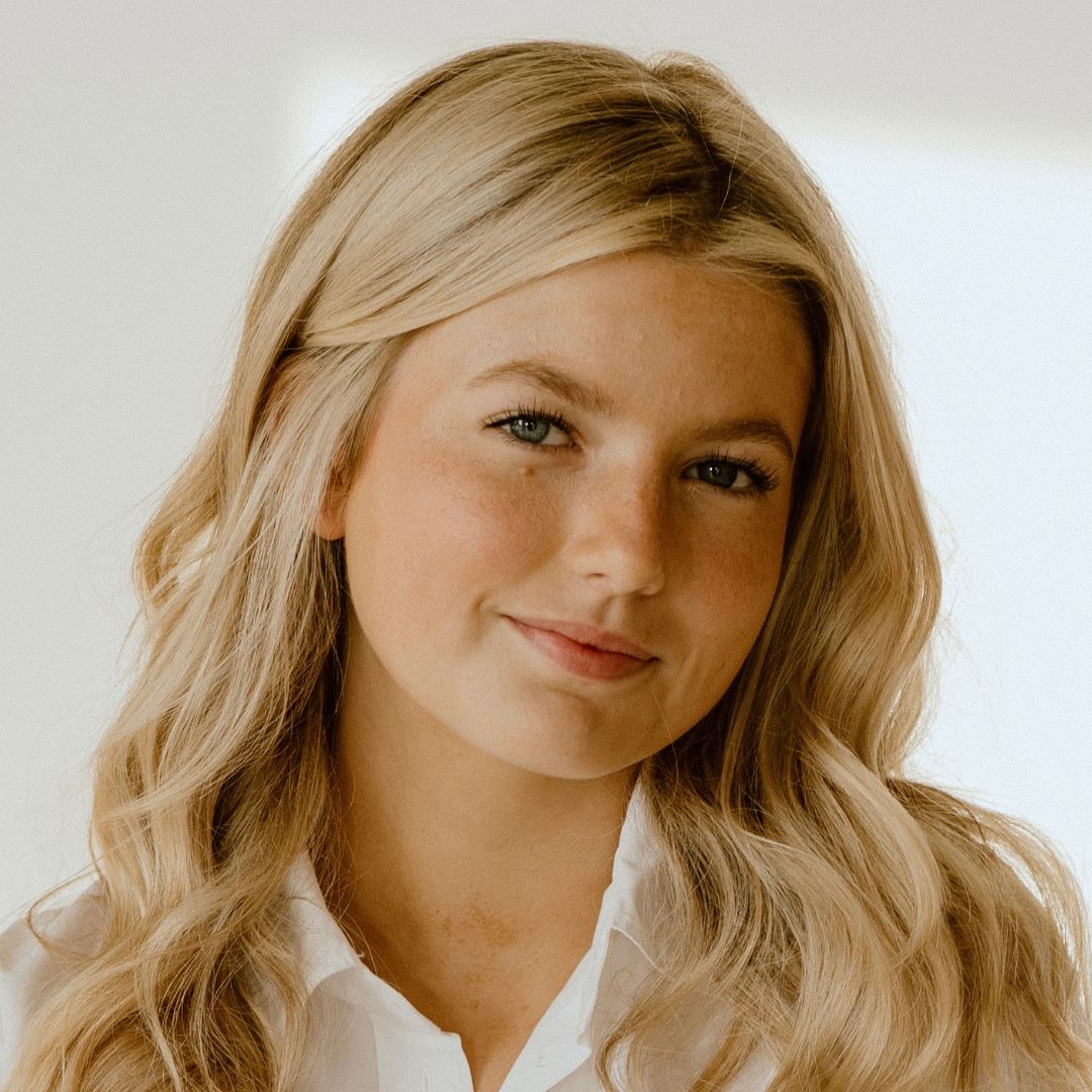 Young woman with blonde, wavy hair and blue eyes smiling gently, wearing a white collared shirt.