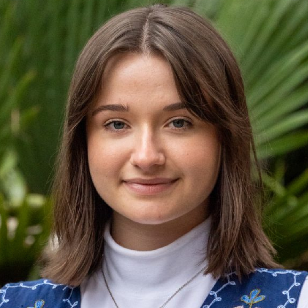 Young woman with light brown hair and blue eyes, smiling subtly, wearing a white turtleneck and a blue embroidered vest.