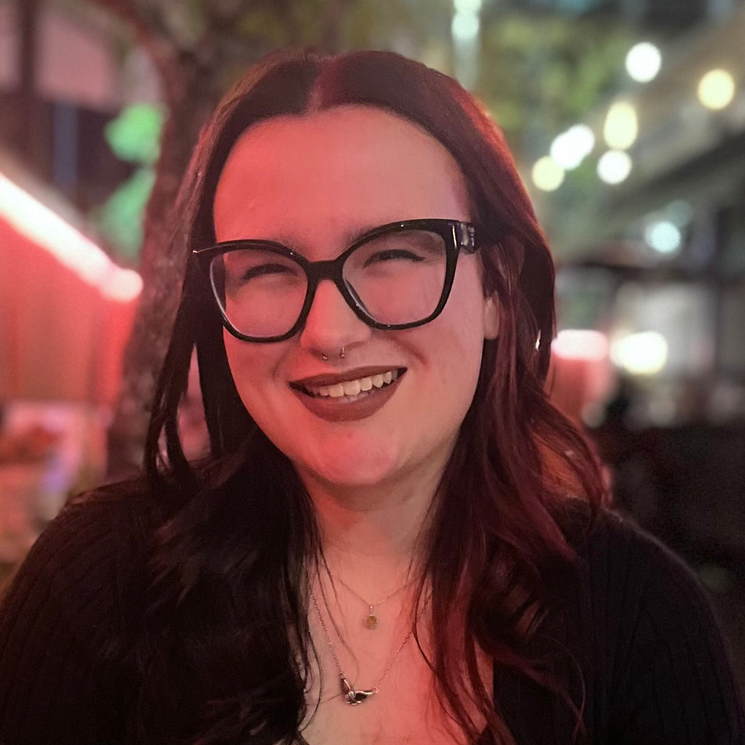 A woman with dark hair and glasses smiles broadly at the camera, with a blurred background of evening lights.