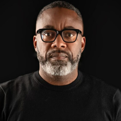 A person with short gray hair, black glasses and a salt-and-pepper beard wears a black shirt against a black background.