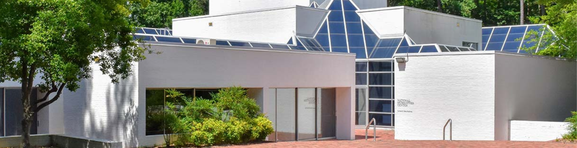 Modern building with geometric design, featuring white walls and large, angular glass windows. Surrounded by trees, it conveys a serene, contemporary feel.