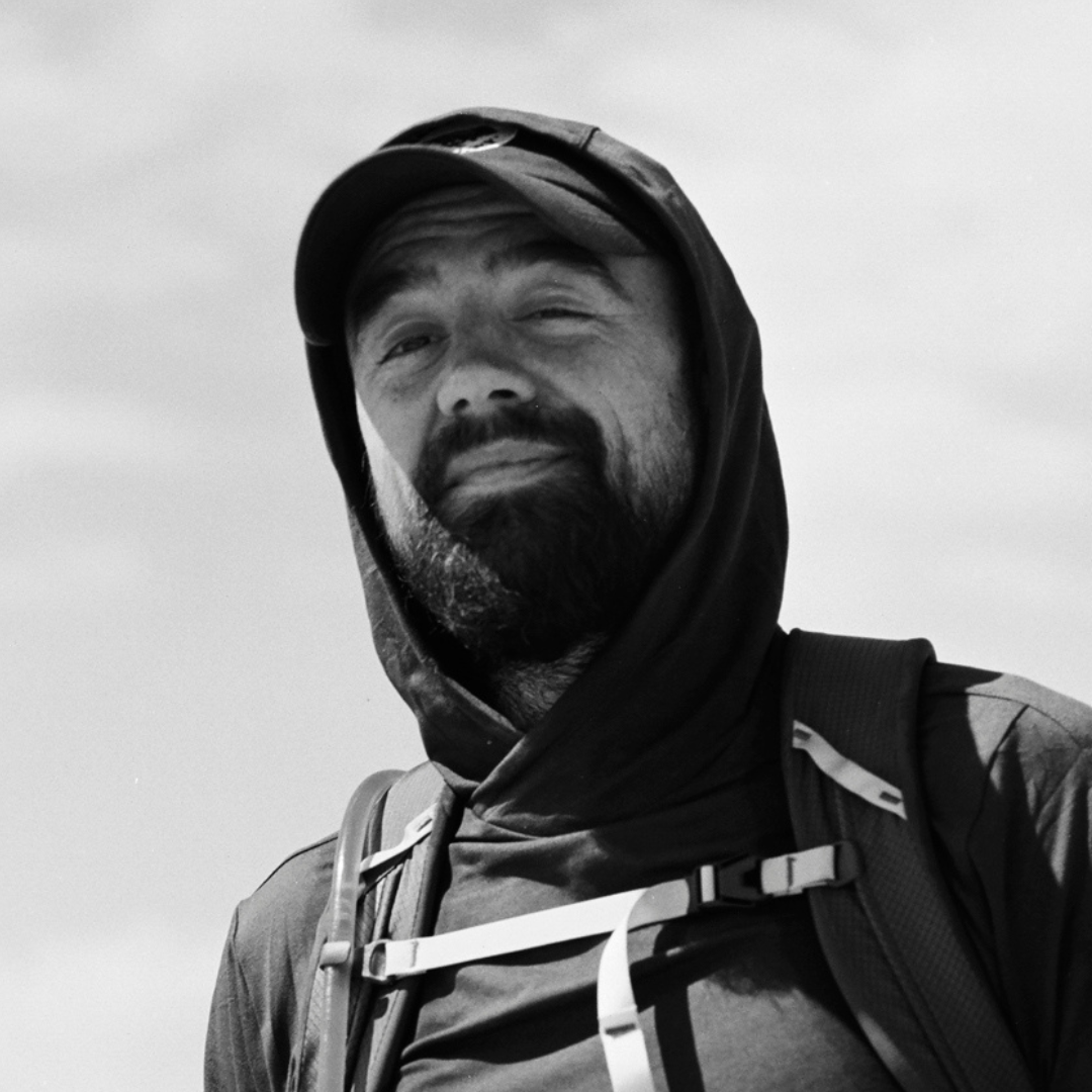 Black and white portrait of a bearded man in a hooded top and baseball cap, looking up with a slight smile, wearing a backpack.