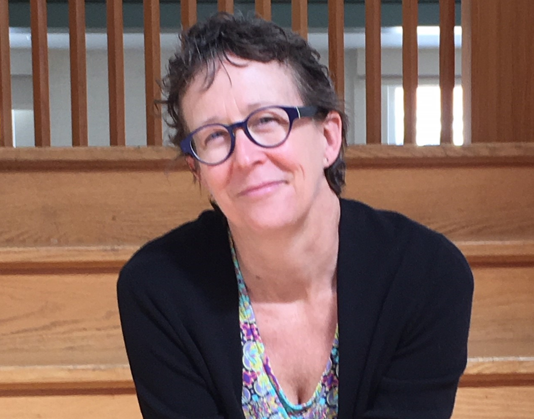 A smiling woman with short, dark, curly hair and glasses, wearing a black cardigan over a colorful patterned top.