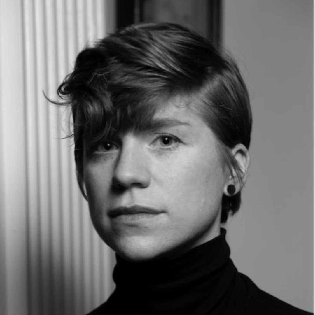 Black and white headshot of a person with short, wavy hair, wearing a black turtleneck and a round earring in their right ear.