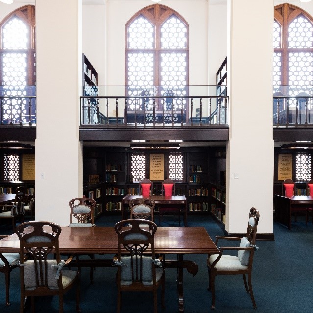 A multi-level library reading room features large arched windows, dark wood paneling, bookshelves, and several wooden tables with chairs, with framed texts displayed on the lower walls.