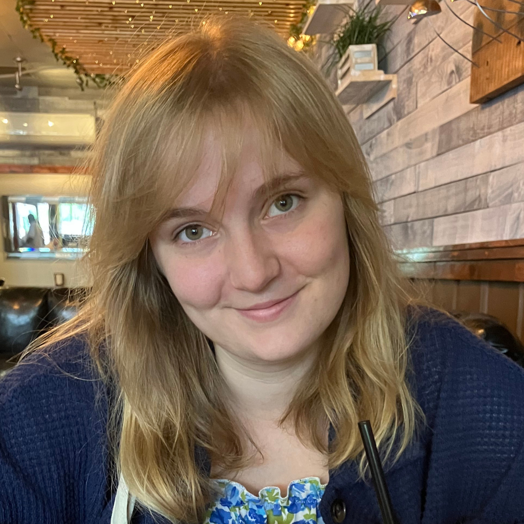 A young woman with blonde hair and green eyes smiles at the camera, wearing a blue cardigan over a floral top in an indoor setting.