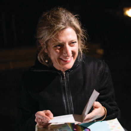 A smiling woman with blonde hair, wearing a black jacket, holds an open card above a colorful gift.