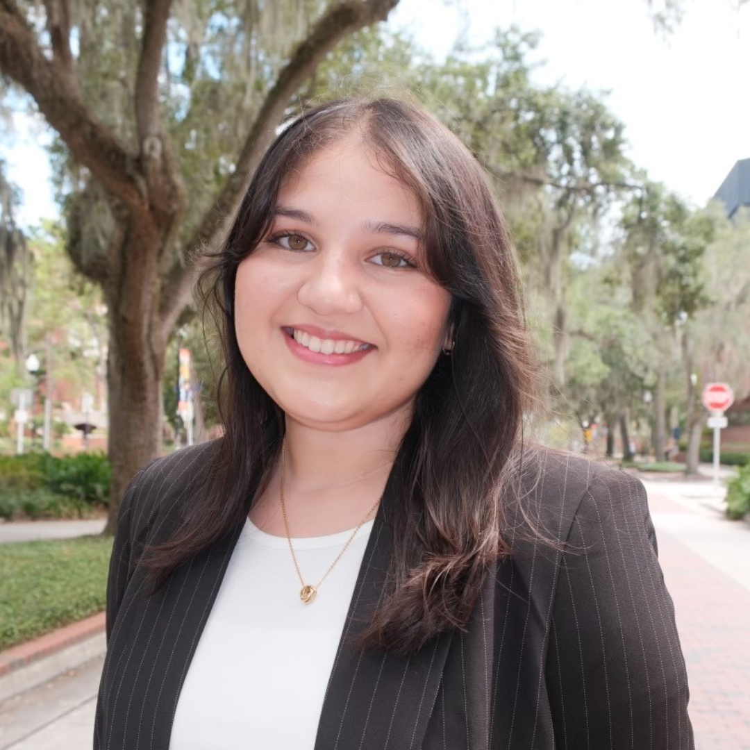 A smiling young woman with dark hair and a pinstripe blazer stands outdoors on a tree-lined street.