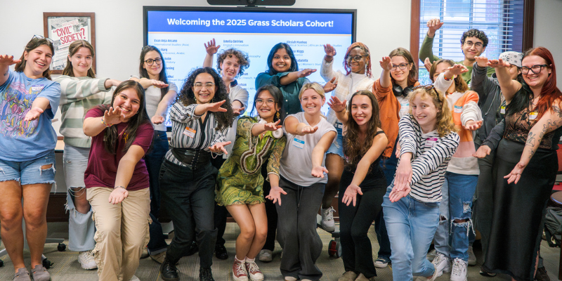 A diverse group of eighteen smiling people poses in a room, standing and kneeling. Behind them, a screen reads "Welcoming the 2025 Grass Scholars Cohort."