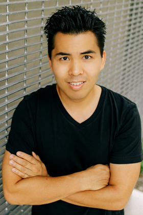 A man with dark hair wearing a black V-neck t-shirt smiles, with his arms crossed, leaning against a metallic grid wall.