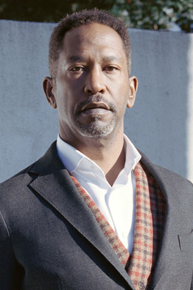 A man with a short beard and dark skin looks directly at the camera, wearing a dark grey blazer, white collared shirt, and a plaid scarf.
