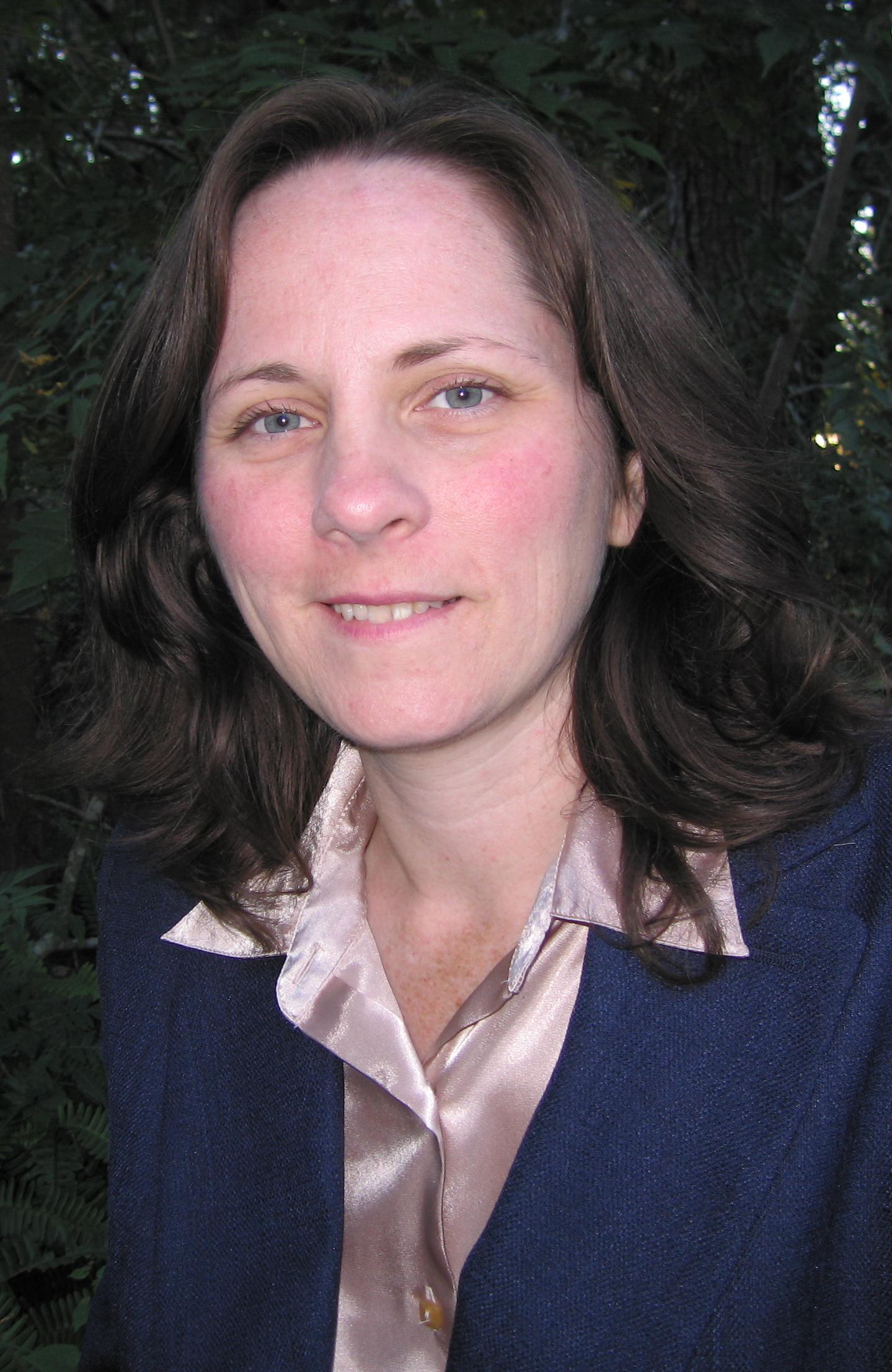 A woman with brown hair and blue eyes smiles at the camera, wearing a blue blazer over a light pink blouse, with a dark green background.