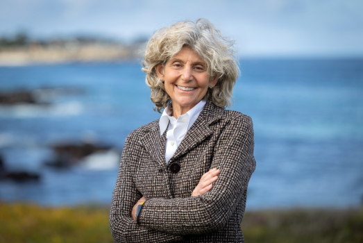 Person with a tweed jacket and button down shirt and gray hair smiling in front of the ocean