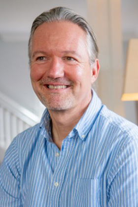 A man with graying hair, wearing a blue and white striped shirt, smiles and looks to his left.