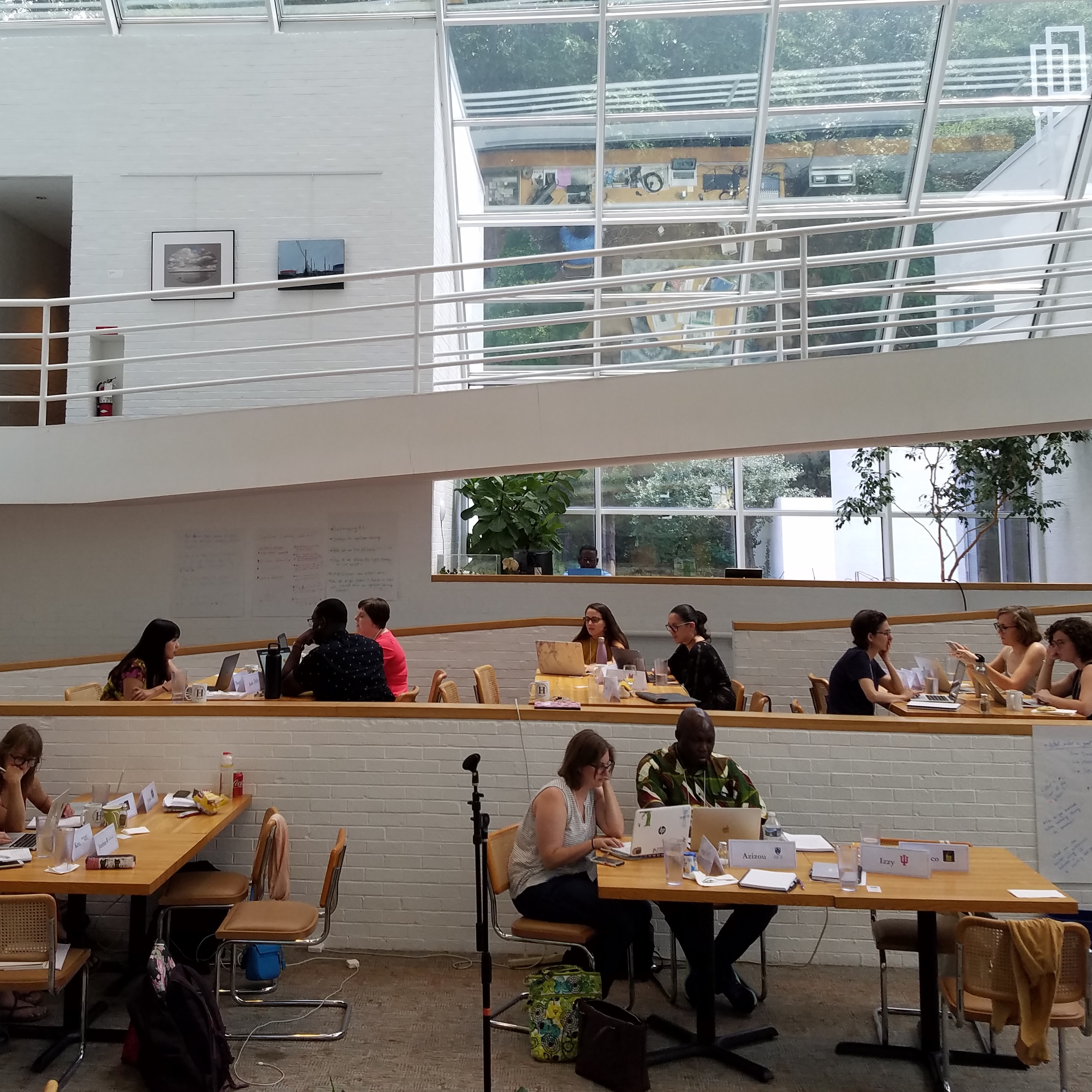 People work on laptops at wooden tables in a bright, modern multi-level space with large windows. Some visible name tags include "Azimou" and "Lizy.